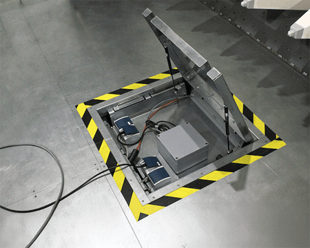 Open floor hatch with exposed electrical components, marked by black and yellow hazard tape