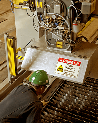 Man in a green hard hat leaning over a machine with a "Danger, Burn Hazard" label on it