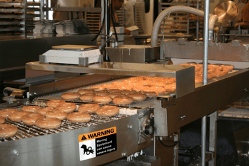Doughnuts moving along a food processing conveyor belt with clear safety labels and signs noting danger.