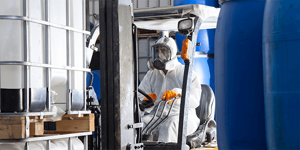 A worker in PPE driving a forklift loaded with chemical storage containers.