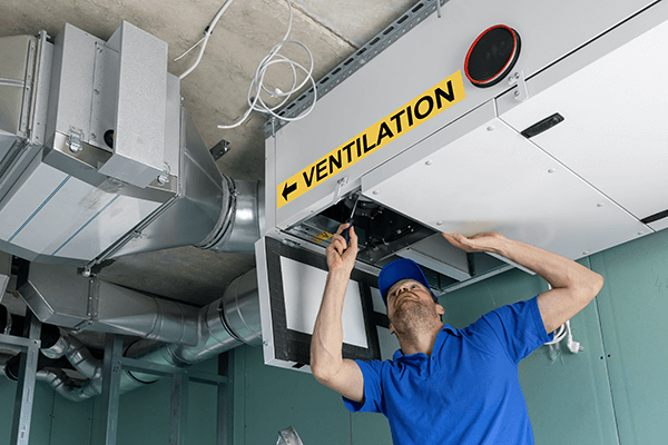  Technician in blue uniform inspecting ceiling ventilation system labeled with yellow ventilation sign