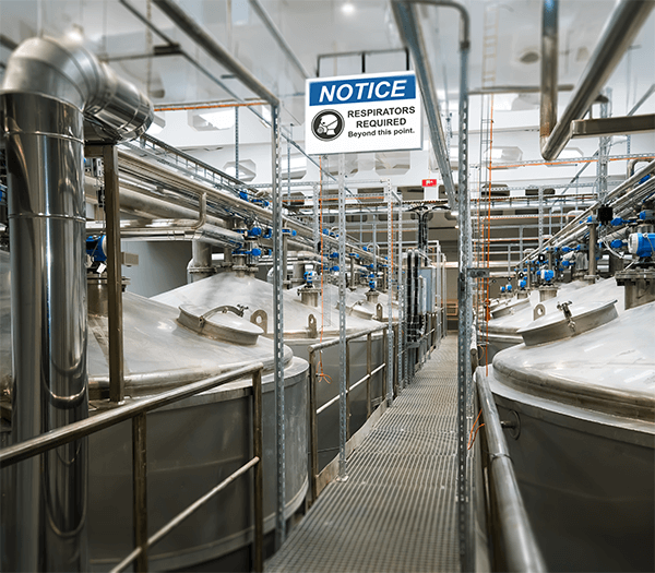  Industrial processing area with metal tanks and overhead sign reading ‘Notice – Respirators Required.’