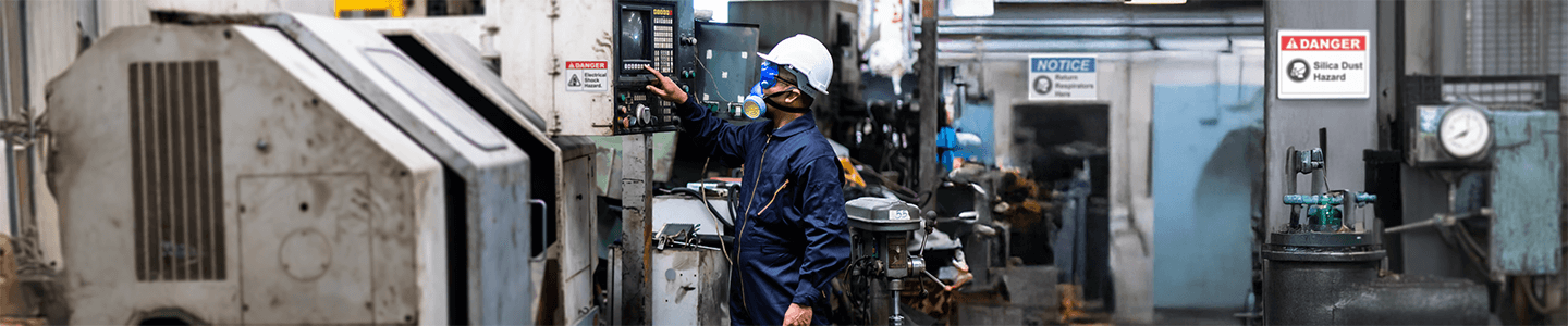 Worker in respirator operating industrial machinery with silica dust hazard and notice signs in background