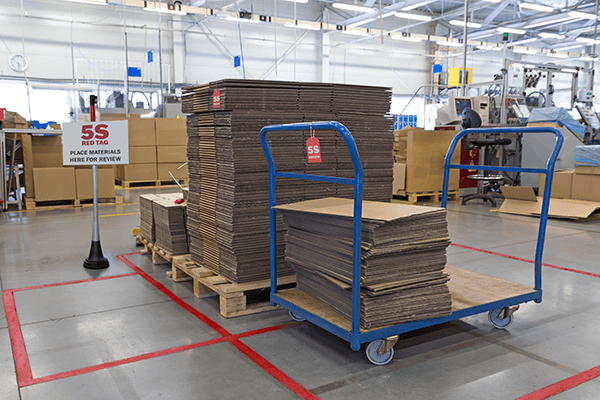 Stacks of cardboard on pallets in a 5S red tag review area marked with red floor tape