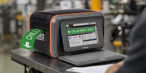 A DuraLabel Toro Max Industrial Label Printer actively printing a green “CHILLED WATER – SUPPLY” pipe marker in a mechanical room workspace. 