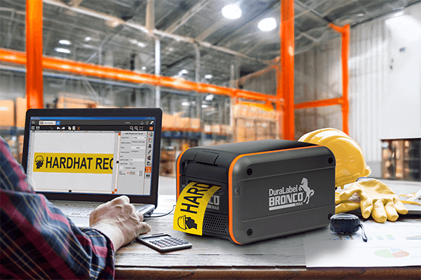 Bronco Max desktop industrial label printer connected to a laptop with LabelForge PRO in a warehouse office.