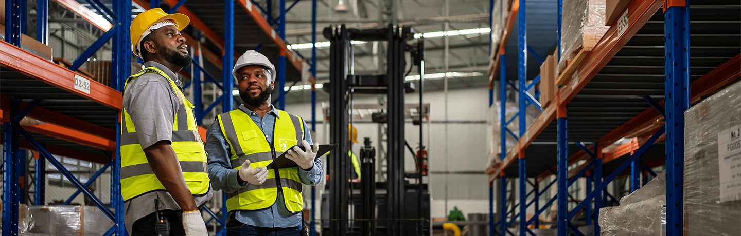 A manager speaking with his crew member standing in a warehouse in front of a forklift.