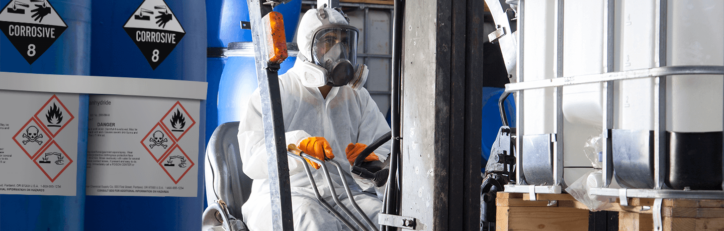 A worker in a Tyvek suit and respirator driving a forklift with HazCom barrels in the foreground