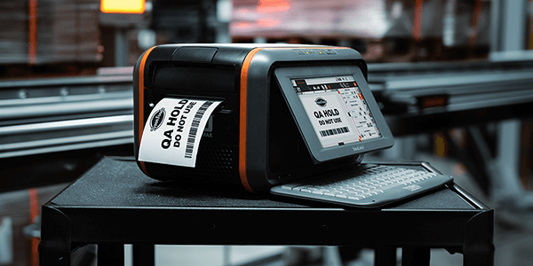 DuraLabel Toro Max printer producing a QA Hold label on a workbench in a warehouse with stacked pallets in the background. 