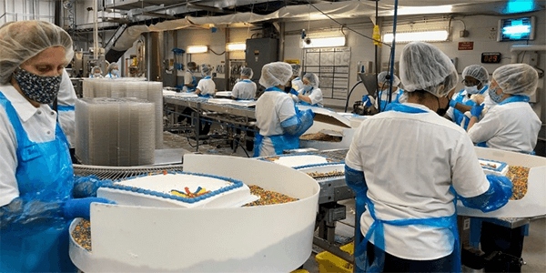 Workers in protective clothing and masks decorate sheet cakes on a food production line inside a Rise Baking Company facility. 