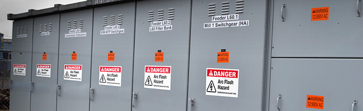 Outdoor electrical switchgear cabinets labeled with warning signs at an industrial facility.