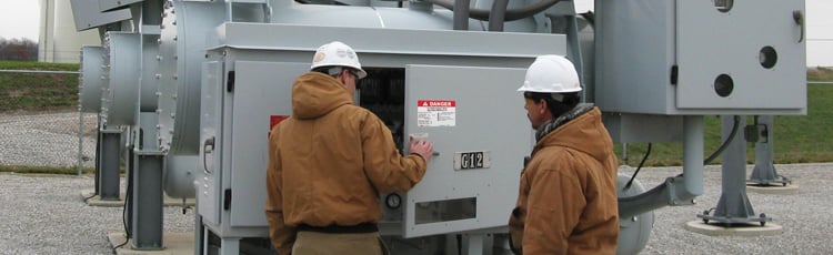 Two workers wearing hard hats and brown jackets inspecting electrical equipment outdoors at an industrial facility.