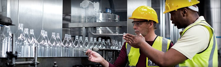 Assembly line workers discuss quality control principles in front of a convey belt transporting glass bottles.