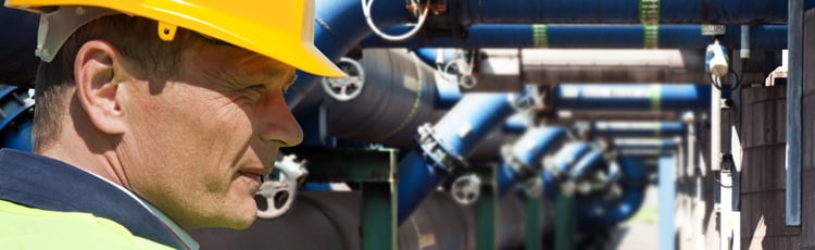 A worker in a yellow hardhat stands in front of an array of pipes and valves.