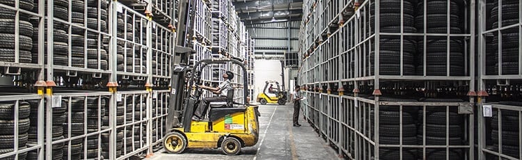 A forklift loads tires on racks in a warehouse with labels and floor lines visible.