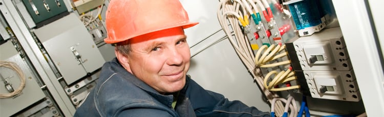 Industrial electrician in an orange hard hat servicing an electrical control panel with exposed wiring for workplace electrical safety compliance.