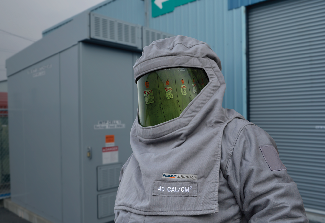 Worker wearing an arc flash protective suit and face shield near electrical equipment.